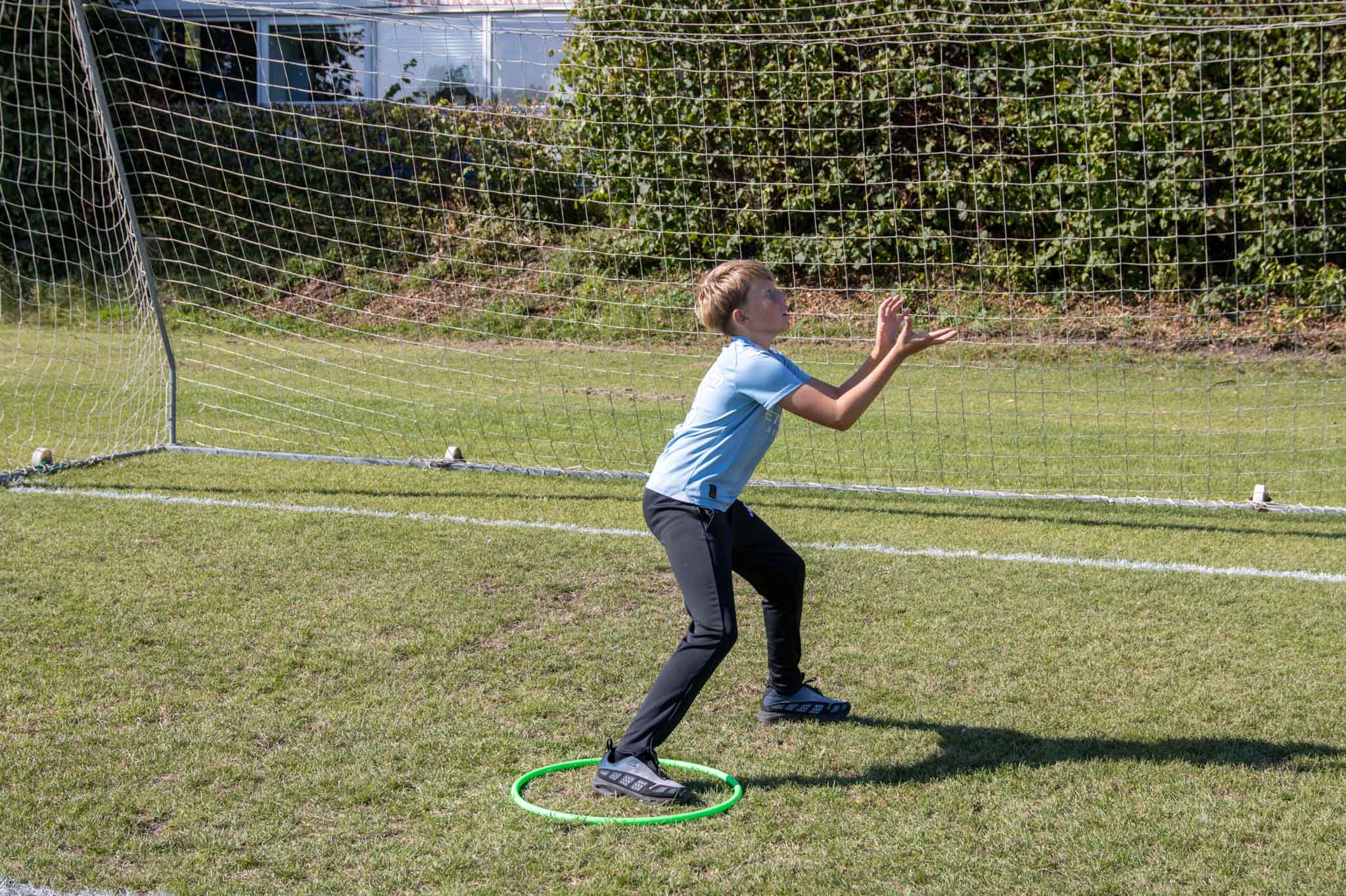 En dreng står i en grøn hulahopring på en græsplæne og gør sig klar til at gribe en bold foran et fodboldmål.